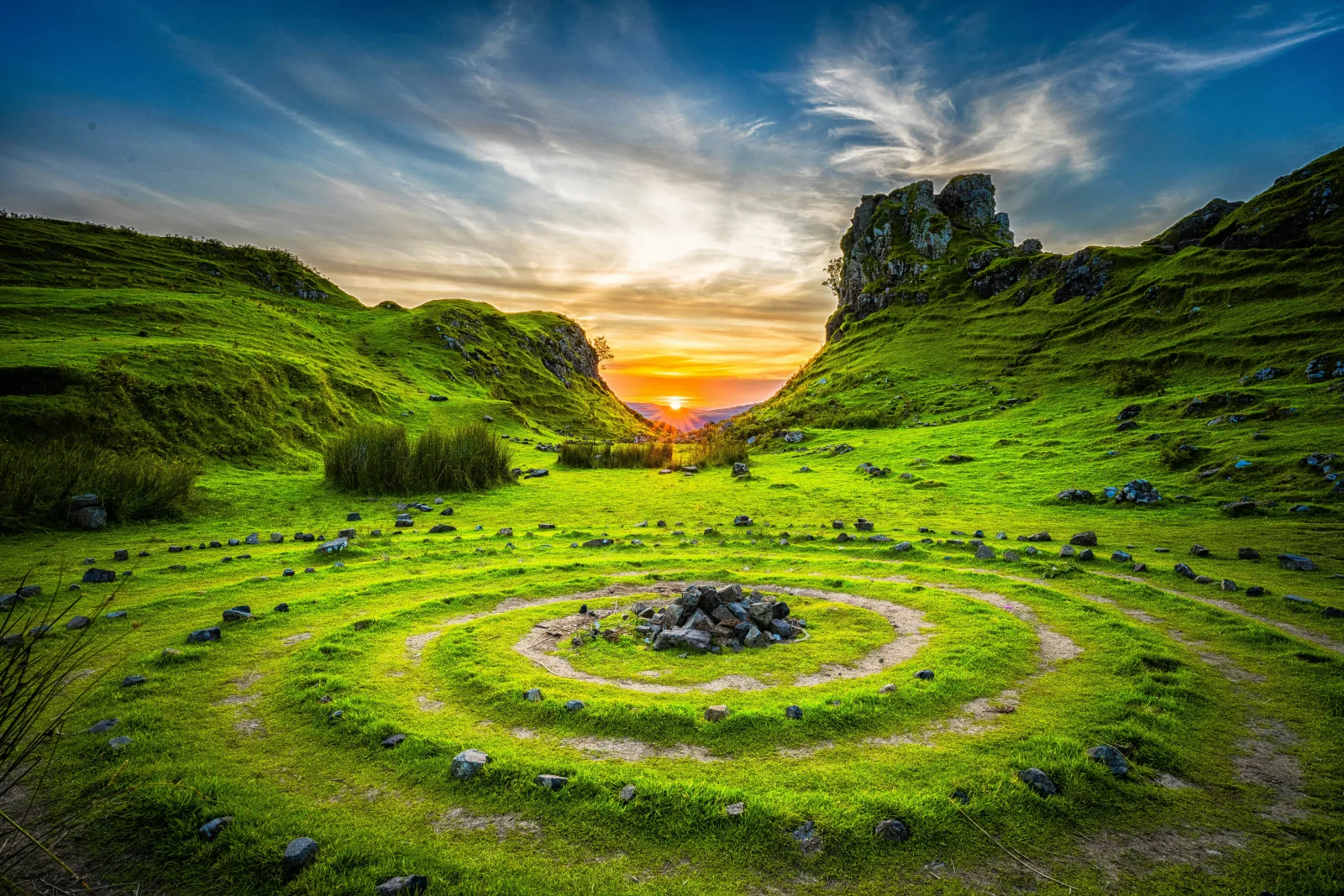 Círculos de pedra desenhados na grama de um vale verde, com o sol se pondo entre os morros
