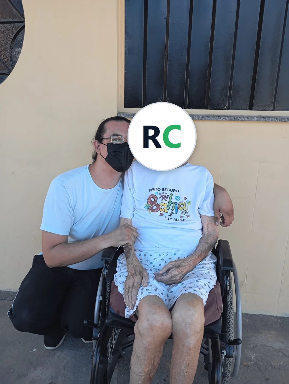 Fisioterapeuta agachado ao lado de uma idosa sentada em cadeira de rodas, em frente a uma casa