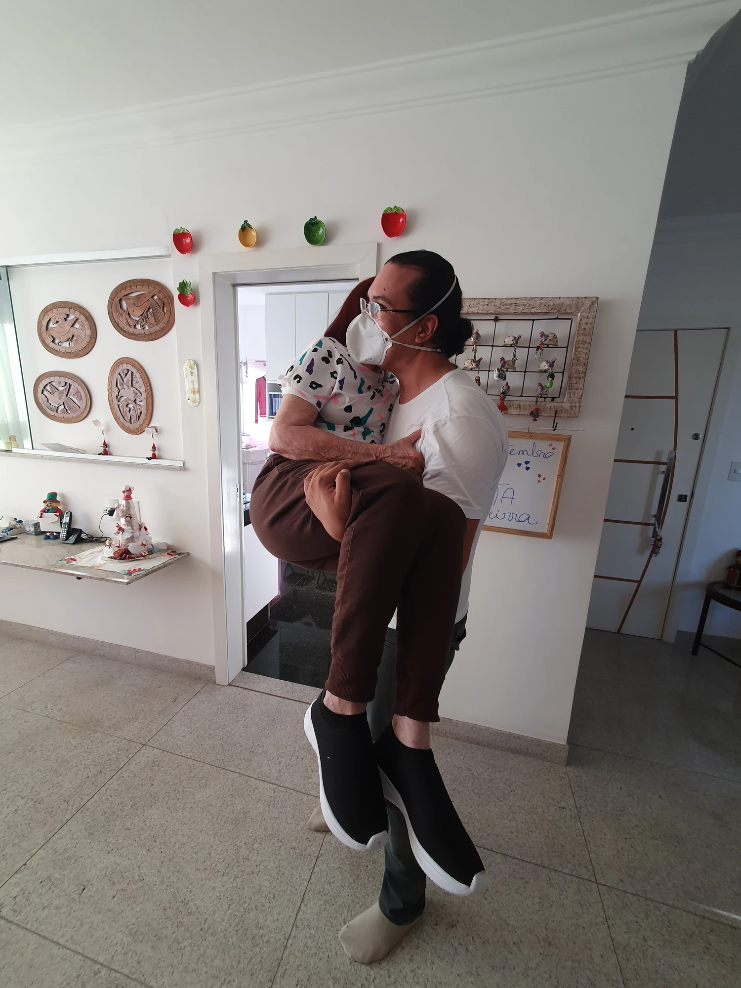 Fisioterapeuta carregando uma idosa no colo dentro de um apartamento